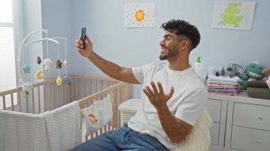 Genç İspanyol bir adam, oyuncaklarla çevrili bir bebek beşiği ile rahat bir yatak odasında otururken telefonuyla video konferans yapıyor..