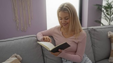 Kadın, rahat bir oturma odasında, rahat bir şekilde otururken kitap okuyor, yumuşak mobilyalarla çevrili, evinde huzurlu bir anın tadını çıkarıyor..