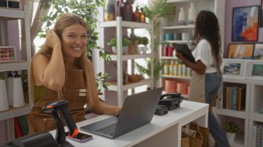 Ev dekorasyonunda çalışan bir kadın dizüstü bilgisayar ve meslektaşı ile arka planda çalışıyor rafları düzenliyor, canlı ve modern bir perakende ortamı sergiliyor.