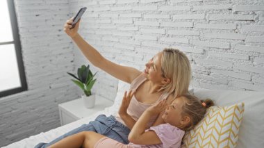 Kadın ve kızı rahat bir yatak odasında, gülümseyerek ve el sallayarak bir aile anını görüntülüyorlar..