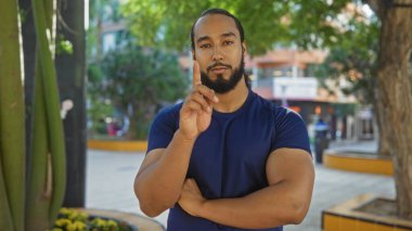 Mavi tişörtlü genç adam şehir parkında duruyor. Arka planda ağaçlar ve binalar bulanık. Düşünceli bir ifadeyle yukarıya doğru bakıyor..