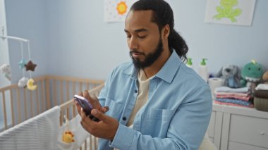 Yatak odasında, bebek beşiğinin yanında akıllı telefon tutan, peluş oyuncaklar ve katlanmış giysilerle çevrili, rahat ve ilgili bir ev ortamını ima eden bir adam var..