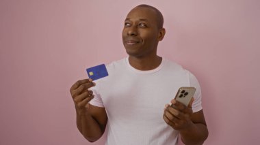 Akıllı telefon ve kredi kartı tutan genç bir adam, modern yaşam tarzı ve teknoloji kullanımını gösteren izole edilmiş pembe bir geçmişe güvenle karşı çıkıyor..