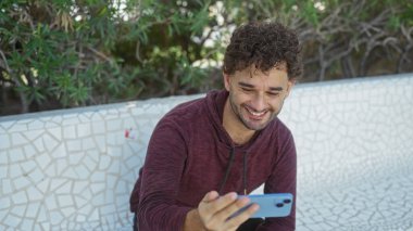 Çevresi yeşil yapraklarla çevrili şehir parkındaki mozaik bankta akıllı telefon kullanırken gülümseyen yakışıklı adam rahat ve modern bir yaşam tarzını aktarıyor.
