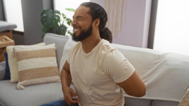 Oturma odasındaki koltukta oturan sakallı genç adam, sırt ağrısı çekiyor, doğal dekor ve iç mekan bitkileriyle, rahat bir dairede günlük beyaz tişört giyiyor..