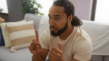 Modern dairenin oturma odasında oturan sakallı genç adam kapalı alandaki çizgili minderlerin yanındaki beyaz koltukta nötr bir yüz ifadesiyle konuşuyor.