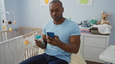 Yatak odasında beşikteki telefonu tutan genç adam, bebek oyuncakları ve temel ihtiyaçlarla çevrili, bu da evde modern bir ebeveynlik sahnesi olduğunu gösteriyor..
