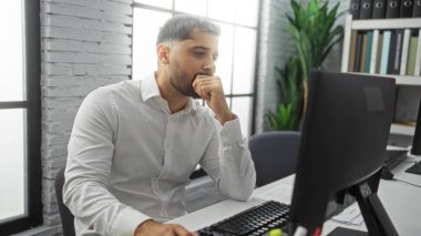 Modern bir ofis ortamında çalışan genç İspanyol bir adam, masa başında oturmuş, parlak bir iç mekanda bilgisayar ekranına odaklanmış, profesyonellik ve konsantrasyon yayıyor..