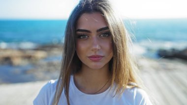 Kumral saçlı genç bir kadın deniz kenarında manzaralı bir gezinti alanında duruyor. Çekici özelliklerini açık bir fon olarak okyanusla sergiliyor..