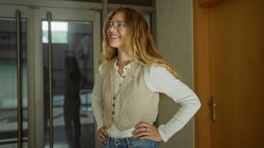 Ofiste kendine güvenen bir kadın, sarı saçlı ve günlük kıyafetleriyle camdan bir arka fona yaslanmış, pozitif ve profesyonel bir atmosferi somutlaştırıyor..