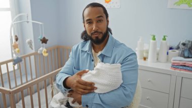 Sakallı genç bir adam, yumuşak pastel renkler ve bebek özleriyle çevrili, sıcak ve huzurlu bir yatak odasında bir bebeği sevgiyle tutuyor..