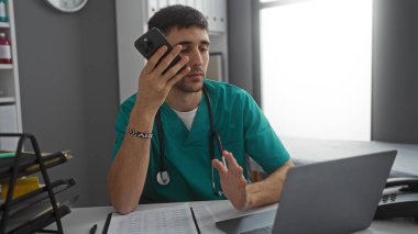 Klinikteki genç İspanyol doktor, bilgisayarın yanında steteskopla iş günü masasında doküman ve boyuna işaret ederken akıllı telefona konuşuyor.