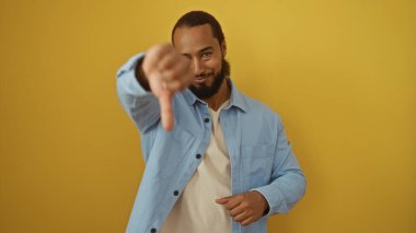 Sakallı genç adam, izole edilmiş sarı arka planı onaylamıyor. Günlük mavi ceket giyiyor ve negatif ifade gösteriyor.