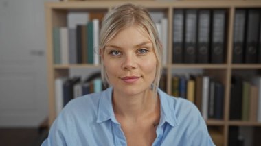 Ofisteki çekici sarışın kadın arka planda kitaplıklarla güvenle gülümsüyor.