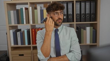 Genç adam bir ofis ortamında cep telefonundan sesli mesaj dinliyor, arka planda kitaplıklar ve dosyalar var, düşünceli ve profesyonel görünüyor.