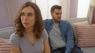 Oturma odasında bir kadın ve bir adam ciddi görünüyor. Ev ortamında bir kanepede oturuyor.