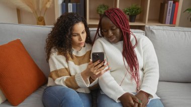 İki kadın, rahat bir oturma odasında, kanepede oturmuş, ev dekoru ve sıcak, davetkar bir atmosferle çevrili akıllı bir telefona bakarken bir anı paylaşıyorlar..