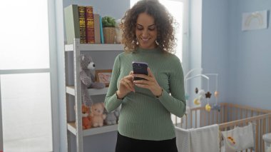 Bebek odasında gülümseyen, akıllı telefon kullanan, oyuncak beşiği olan bir kadın, oyuncak ayılarla dolu bir kitaplığın yanında,
