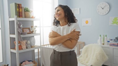 Sıcak bebek odasında gülümseyen bir kadın, kolları çapraz, beşikte duruyor, doldurulmuş hayvanlarla çevrili, günlük kıyafetler giyiyor, doğal ışık ve yumuşak renkte huzurlu bir ortam.