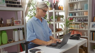 Dizüstü bilgisayar mağazasında çalışan kadının rafları çeşitli dekorasyonlar, güzellik ürünleri ve bitkilerle dolu..
