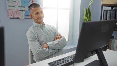 Kolları çapraz bir şekilde ofiste oturan bir adam parlak ışıklı bir iş yerinde arka planda bir bitki ve notlarla bir bilgisayar ekranına bakıyor.