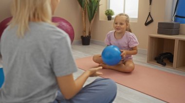 Kadın ve çocuk, kapalı spor salonunda yoga minderleri üzerinde top oynuyorlar. Spor yaparken aile bağı kuruyorlar.