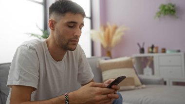 Kanepede oturan genç adam rahat bir oturma odasındaki akıllı telefonu kullanarak modern iç tasarım, pastel duvarlar ve kapalı alan bitkilerine sahip..