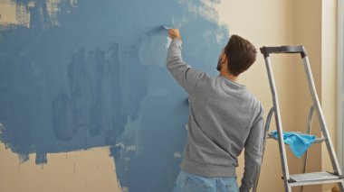 Genç adam yeni evin içini mavi duvarla boyuyor, merdiven, günlük gömlek ve kot pantolon ile kapalı alanda çalışıyor, yaratıcı bir şekilde konforlu apartman alanını yenilemeye odaklanıyor..