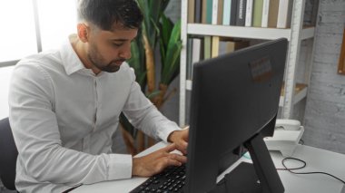 Beyaz gömlekli genç adam modern ofis alanında bilgisayar başında çalışıyor. Kapalı mekanlar ve kitaplıklar ile profesyonel bir ortamda görevine odaklanıyor..