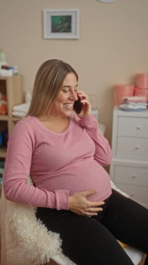 Genç hamile bir kadın yatak odasında oturuyor telefonda konuşuyor. Ev içinde rahat bir yerde, beşiğin yanında..