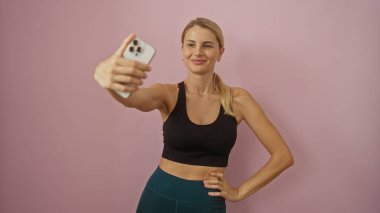 Pembe arka planda gülümseyen, siyah spor tişörtlü, saçları omuzuna bağlanmış, kendinden emin ve rahat bir şekilde selfie çeken bir kadın..