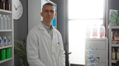 Eczanede duran genç adam beyaz laboratuvar önlüğü giyiyor. Etrafı bardaklar ve şişeler de dahil olmak üzere çeşitli tıbbi ve sağlık ürünleriyle dolu raflarla çevrili.