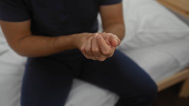 Yatak odasında bir yatakta oturan genç adam, ağrıyan bileğini tutuyor, rahatsız olduğunu gösteriyor, odaklanmış bir ifadeyle, bir ev ortamında acı ve iç gözlem anı yakalıyor..