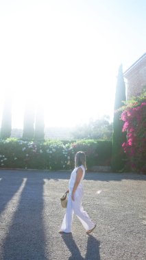 Genç bir kadın güneşli Mallorca, İspanya 'da yürüyor, beyaz elbiseler giyiyor ve adada yemyeşil ve çiçeklerle çevrili saman bir çanta taşıyor.