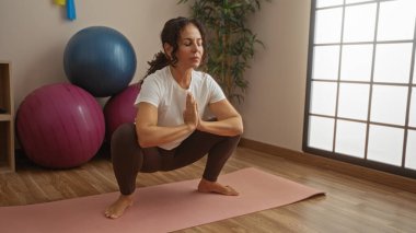 Spor merkezinde yoga yapan bir kadın, egzersiz ekipmanlarıyla çevrili, dinginlik taşıyan ve kendini spora adamış bir spor salonuna odaklanmış..