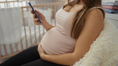 Akıllı telefonlu hamile bir kadın rahat bir bebek odasında karnına dokunuyor yumuşak battaniyelerle süslenmiş tahta bir beşiğin yanında huzurlu bir ev ortamı yaratıyor..