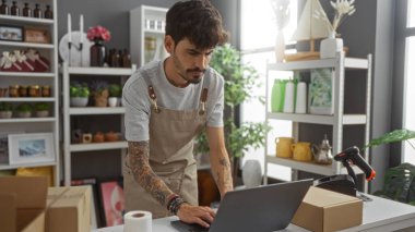 Ev dekorasyonu mağazasında çalışan genç adam dizüstü bilgisayarı ve odaklanmış ifadesiyle güzel organize edilmiş raflar ve dekoratif eşyalarla çevrili..