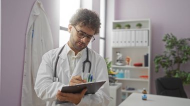 Gözlüklü, beyaz önlüklü, profesyonel ekipman ve bilgilendirici malzemelerle çevrili, iyi düzenlenmiş bir tıp ofisinde pano okuyan genç bir adam..
