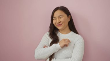 Beyaz kazaklı, gülümseyen genç Çinli bir kadın uzun saçlı, pembe bir arka plana karşı güvenle duruyor ve kolları katlanmış, güzelliği ve özgüveni resmediyor..