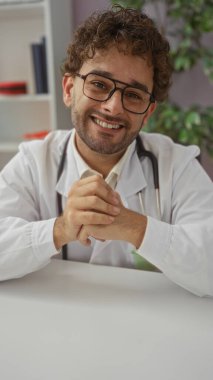 Klinikteki işyerinde gülümseyen bir doktor, güvenle oturur, gözlük takar, steteskop takar ve profesyonel bir portrede, katlanmış eller ve dostane bir ifade takınır..