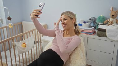 Yatak odasında rahat bir şekilde selfie çeken bir kadın yanında bebek beşiği olan ve rahat bir ev dekorunda mutlu bir şekilde gülümseyen, rahat ve neşeli bir iç mekan anı gösteren bir kadın..