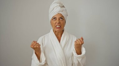 Beyaz bornoz ve havlu giymiş kızgın bir kadın, izole edilmiş beyaz arka planda hayal kırıklığını gösteriyor, yumruklarını sıkmış ve yoğun bir ifadeyle.