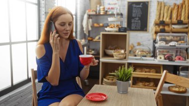 Genç bir kadın elinde kırmızı bir kahve fincanı tutarken telefonda konuşuyor. Fırının içinde çeşitli hamur işleriyle birlikte..