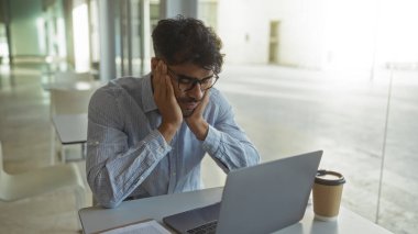 Klinik masasında dizüstü bilgisayarda çalışan sakallı ve gözlüklü genç adam elleri yüzü asık, boş sandalyeler ve kahve fincanıyla çevrili..