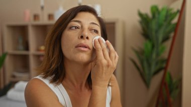 Middle-aged woman applying facial treatment in a spa with a cotton pad for skin care in an indoor wellness center clipart