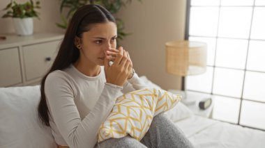 Genç, çekici esmer kadın yatak odasında bir yatakta oturuyor, sıcak bir evde yastığını tutarken bir bardaktan içiyor..