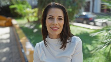 Şehir parkında gülümseyen, esmer saçlı, güneş ışığının altında beyaz bluz giyen, hayat dolu şehir manzarasında güzellik ve sükunet gösteren bir kadın..