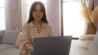 Genç Asyalı kadın, Çin 'de rahat bir oturma odasında dizüstü bilgisayarla çalışırken gülümsüyor..