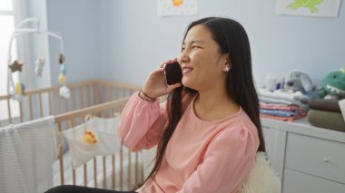 Bebek odasında beşikte telefonla konuşan, pembe bluz giyen, gülümseyen, rahat ev ortamında genç ve çekici Asyalı bir kadın..