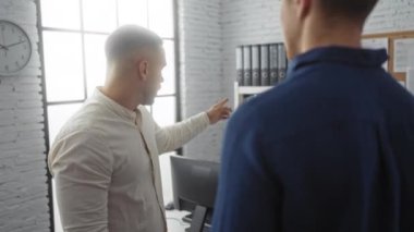 Modern bir ofiste çalışmayı tartışan adamlar, rafların yanında duran halka klasörleri ve beyaz tuğla duvarda bir saat, işbirlikçi ve profesyonel bir iç mekan yaratıyorlar.